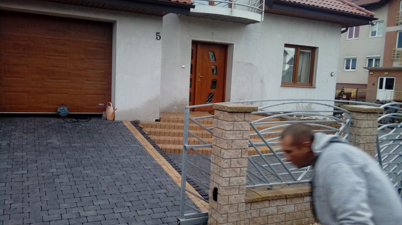 Ułożona szara kostka brukowa przed domem w Bydgoszczy, z widocznym fragmentem nowo zamontowanego ogrodzenia i schodów wejściowych.
