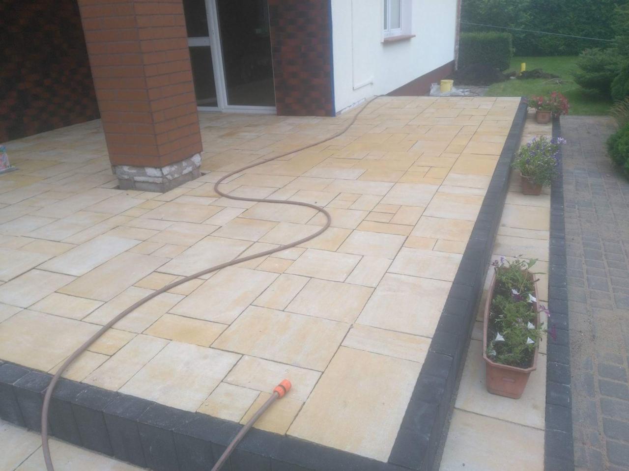 Wykończony taras z jasnej kostki brukowej o nieregularnym kształcie, obrzeża z ciemnej kostki, widoczny fragment domu z ceglanym filarem i wąż ogrodowy na powierzchni.