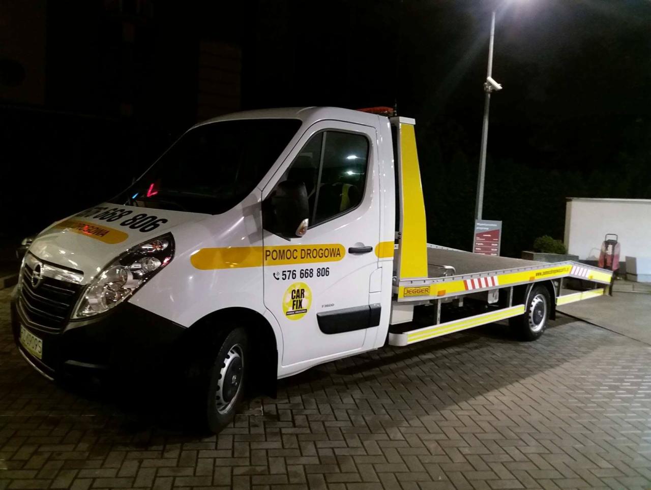Biała laweta Opel z żółtymi elementami i napisem 'Pomoc Drogowa' oraz numerem telefonu, zaparkowana na brukowanej powierzchni nocą pod latarnią.