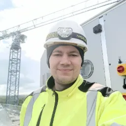 Mężczyzna w kasku z lampką czołową i żółtej kurtce roboczej, na tle dźwigu budowlanego i panelu sterowania z przyciskami.