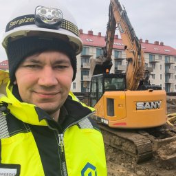 Mateusz Porębski - Uśmiechnięty inżynier w kasku z latarką na tle żółtej koparki SANY ST1550 i budynku mieszkalnego. Widoczny identyfikator firmy.