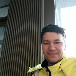 Mateusz Porębski - Mężczyzna w żółtej kurtce na tle grzejnika panelowego i okna. Widoczny fragment instalacji grzewczej w budynku. Ujęcie z bliska, selfie.