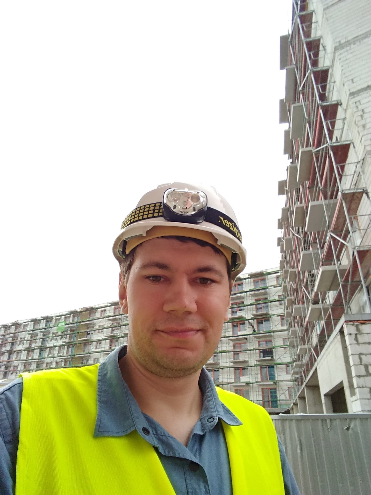 Mężczyzna w kasku z lampką czołową i żółtej kamizelce odblaskowej na tle budynku w budowie z rusztowaniami.