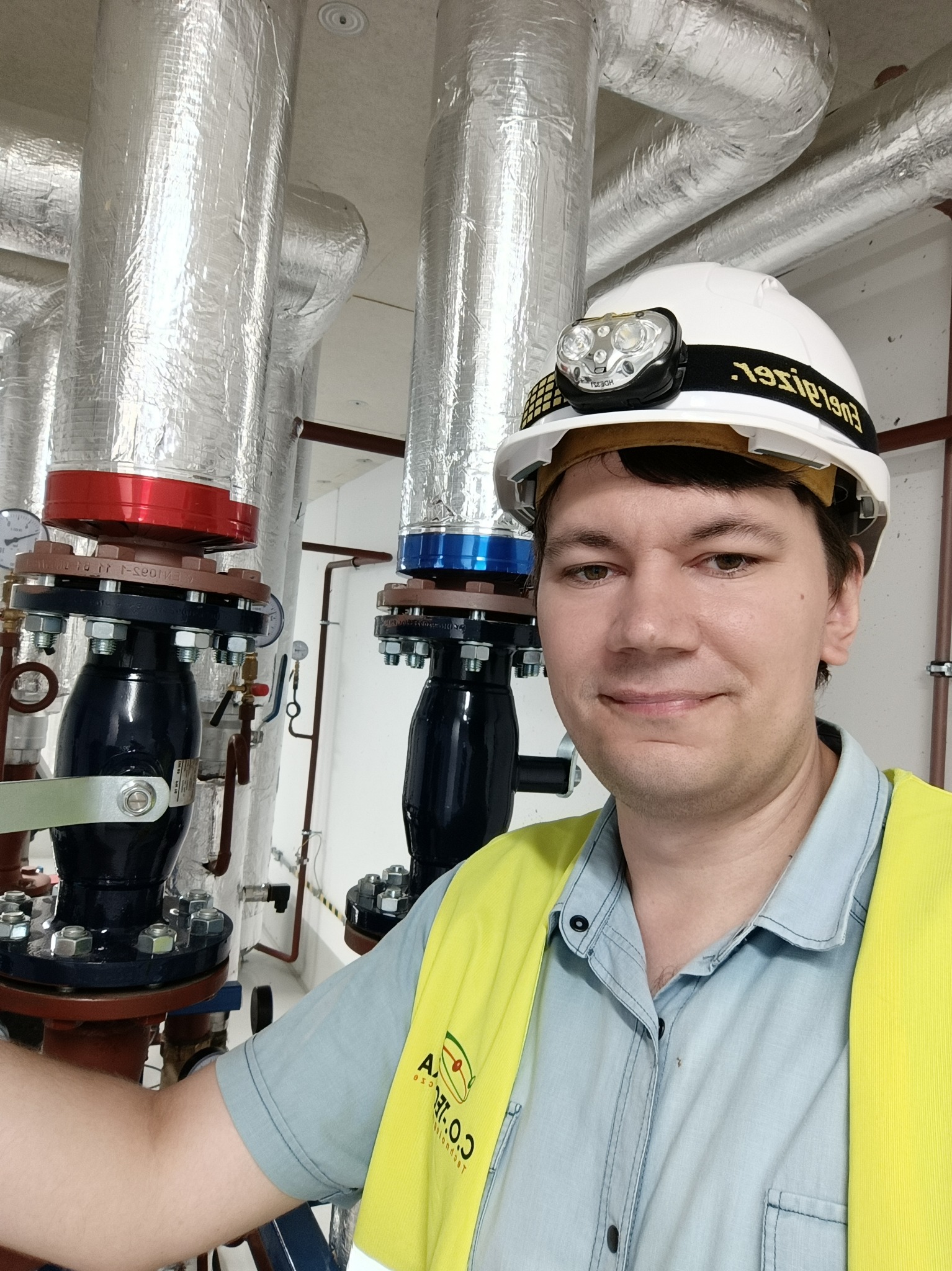 Mężczyzna w kasku z lampą czołową, ubrany w kamizelkę odblaskową, stoi przed rurami z izolacją termiczną i zaworami z manometrami w kotłowni.