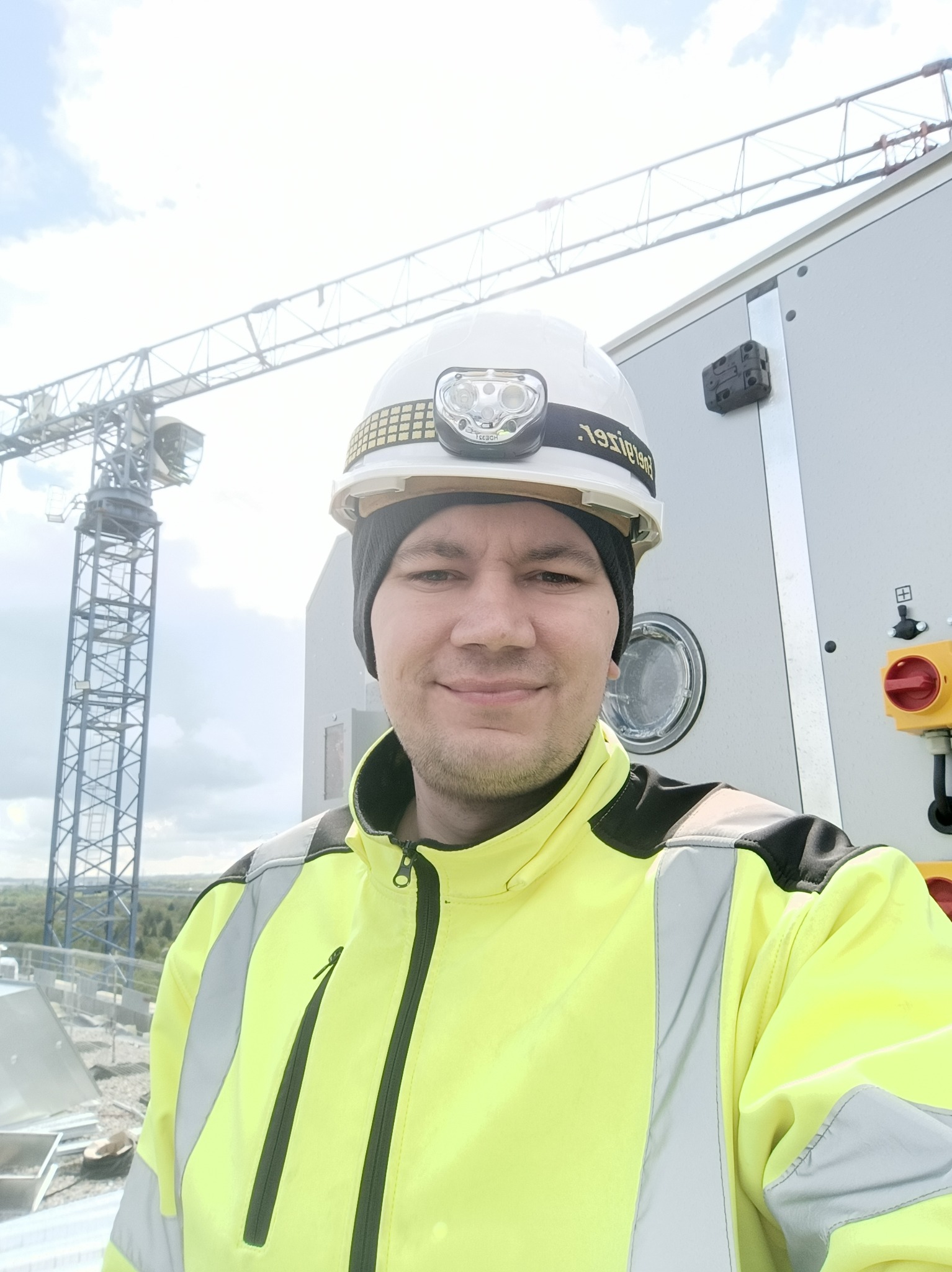 Mężczyzna w kasku z lampką czołową i żółtej kurtce roboczej, na tle dźwigu budowlanego i panelu sterowania z przyciskami.