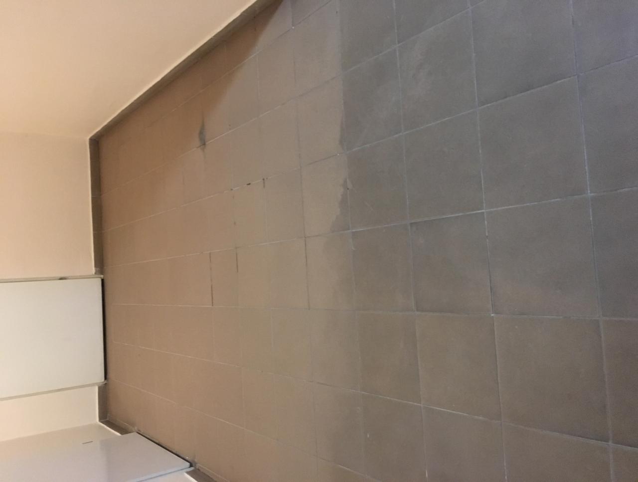 Częściowo umyta ściana z szarych płytek w pomieszczeniu, widoczna różnica w kolorze pomiędzy czystą i brudną powierzchnią, w tle fragment białej szafki.