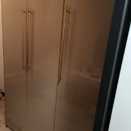 Beżowa, trzydrzwiowa szafa z połyskiem i złotymi, długimi uchwytami, widoczna częściowo w odbiciu toaletka z akcesoriami.