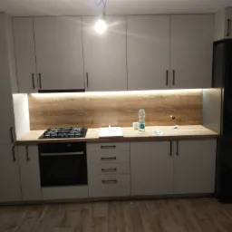 Nowoczesna kuchnia z szarymi szafkami, drewnianym blatem i czarnymi uchwytami, oświetlona punktowo pod szafkami wiszącymi i centralnie zwisającą żarówką.