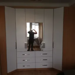 Biała szafa wnękowa z lustrem, widoczne szuflady z poziomymi uchwytami, odbicie osoby robiącej zdjęcie telefonem.