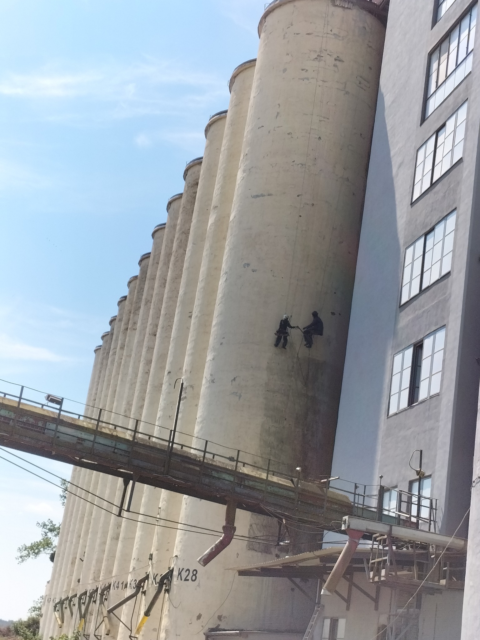 Dwóch pracowników na linach zabezpieczających, wykonujących prace na elewacji wysokiego silosu, widoczny fragment przejścia nadziemnego.