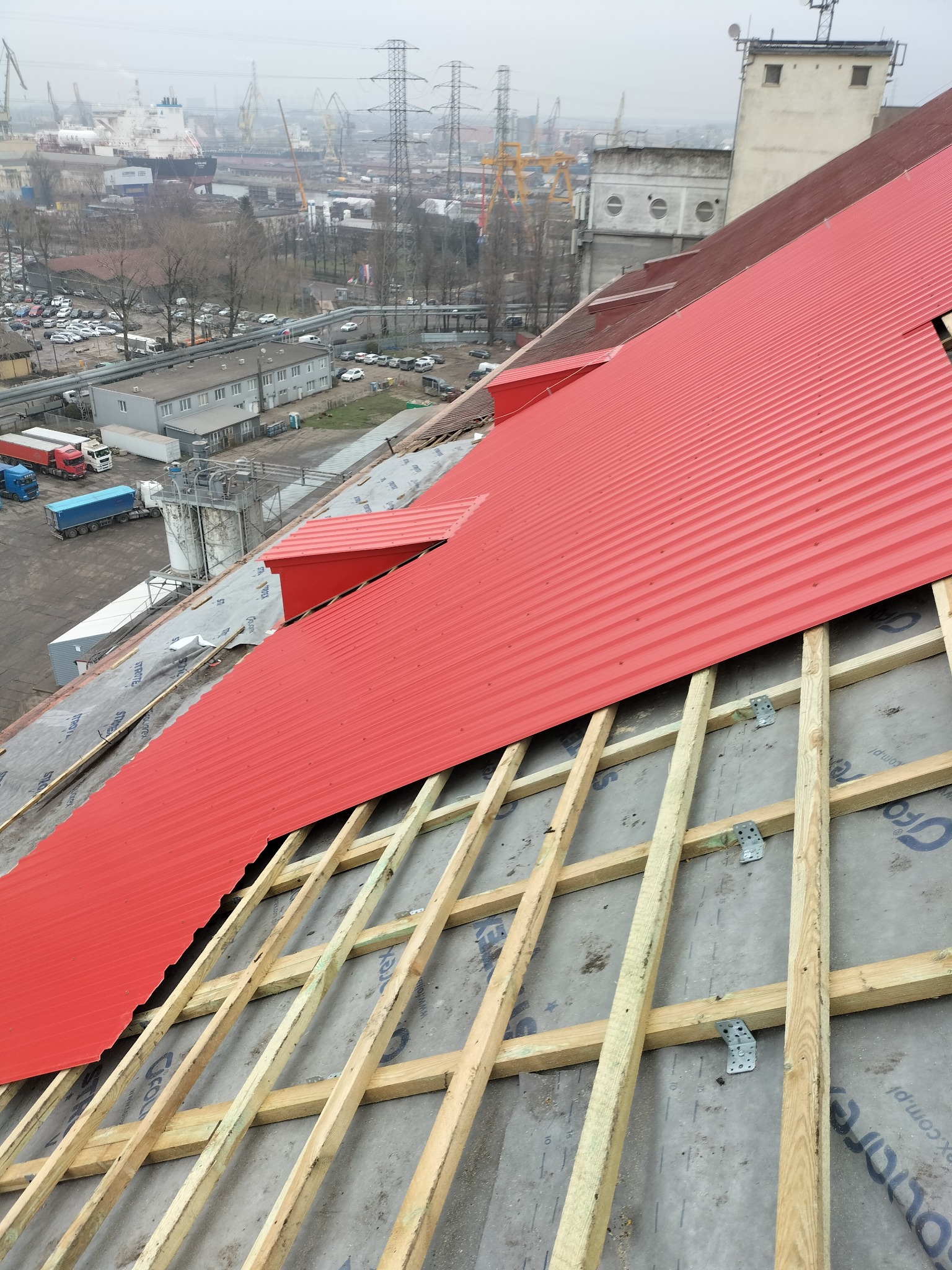 Częściowo pokryty czerwonym blachodachówką dach w trakcie budowy, widoczne drewniane krokwie i membrana dachowa, w tle zabudowania przemysłowe i port.