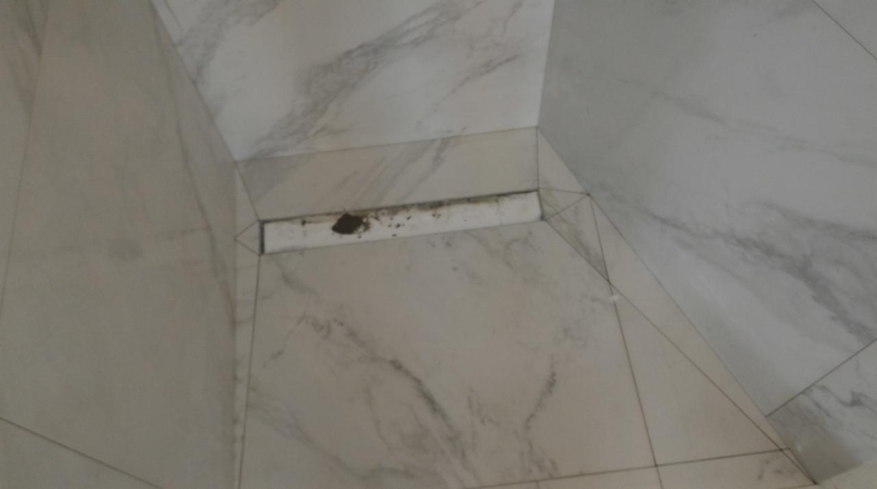 Narożnik kabiny prysznicowej wyłożony białymi płytkami imitującymi marmur; widoczny odpływ liniowy z zabrudzeniami.