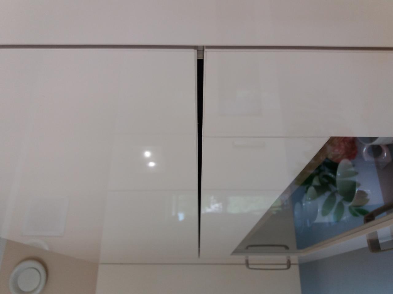 Białe, lakierowane szafki kuchenne podwieszane, z widoczną szczeliną między drzwiczkami i odbiciem blatu w połyskującej powierzchni.