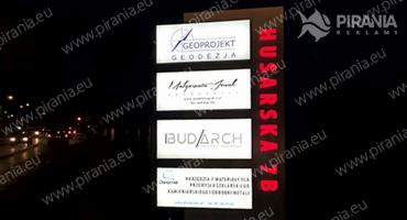 Oświetlony pylon reklamowy z logo różnych firm, umieszczony przy ulicy, widoczny w nocy. Na brązowym tle pionowo umieszczony napis 'HUSARSKA 7B' wykonany z czerwonych, świecących diod.