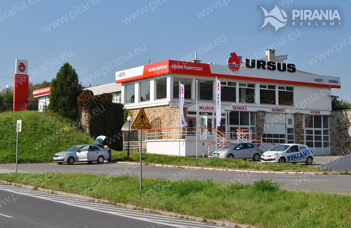 Budynek salonu fabrycznego Ursus z reklamą zewnętrzną, widoczne logo firmy na dachu i na słupie, zaparkowane samochody.