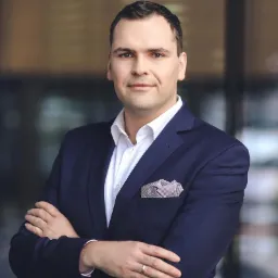 Portret mężczyzny w granatowym garniturze i białej koszuli, z poszetką w kieszonce, stojącego na tle szklanej fasady budynku.