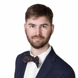 Portret młodego mężczyzny z brodą, ubranego w granatowy garnitur i bordową muchę w białe kropki, uśmiechającego się w kierunku kamery na białym tle.