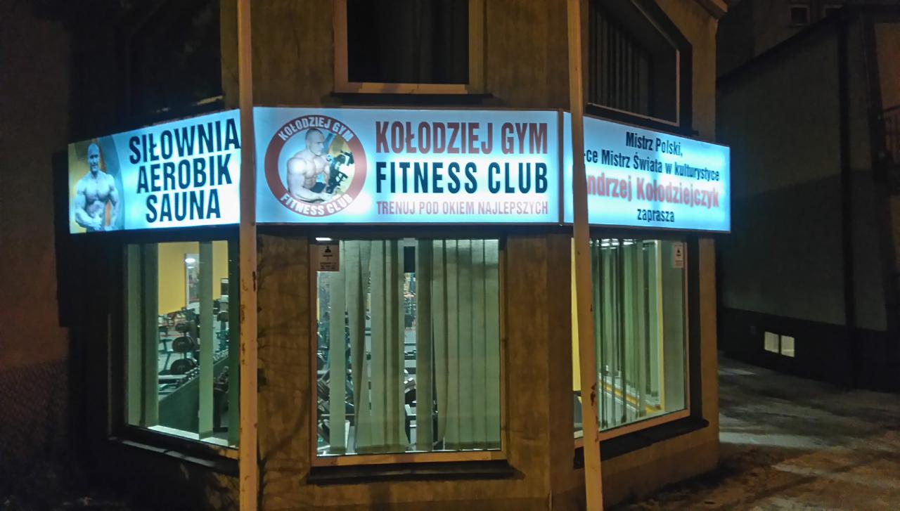 Oświetlony szyld Kołodziej Gym Fitness Club z ofertą: siłownia, aerobik, sauna i zaproszeniem od Andrzeja Kołodziejczyka, Mistrza Polski i Świata w kulturystyce.