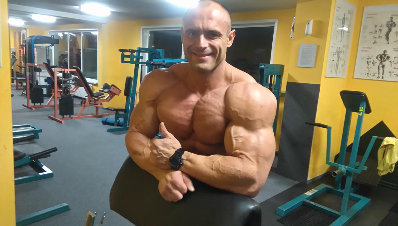 Uśmiechnięty, muskularny mężczyzna w czarnym zegarku, siedzący w siłowni, z uniesionym kciukiem w górę.