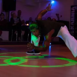 Mężczyzna w białych spodniach i czarnej koszulce wykonuje dynamiczną figurę taneczną na podświetlonej zielonymi laserami podłodze, z widownią w tle.