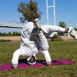Osoba w białym dresie wykonuje stój na głowie na fioletowej macie na trawie, z nogami uniesionymi do góry, w tle most i rzeka.