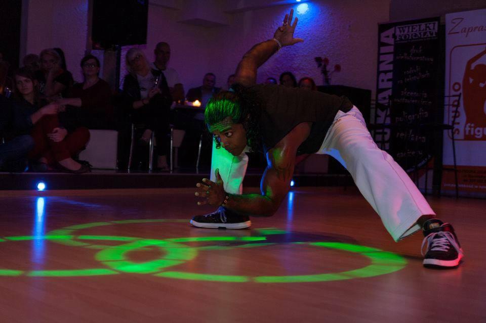 Mężczyzna w białych spodniach i czarnej koszulce wykonuje dynamiczną figurę taneczną na podświetlonej zielonymi laserami podłodze, z widownią w tle.