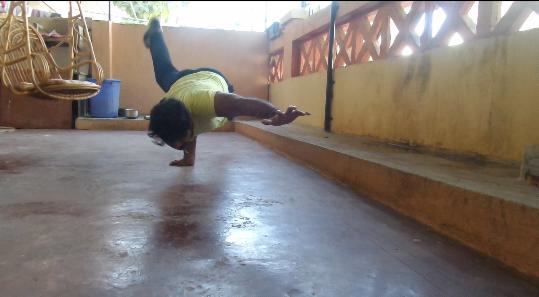 Mężczyzna w żółtej koszulce wykonuje zaawansowaną pozycję odwróconą na jednej ręce na tarasie, z uniesioną nogą i widocznym wiszącym fotelem w tle.