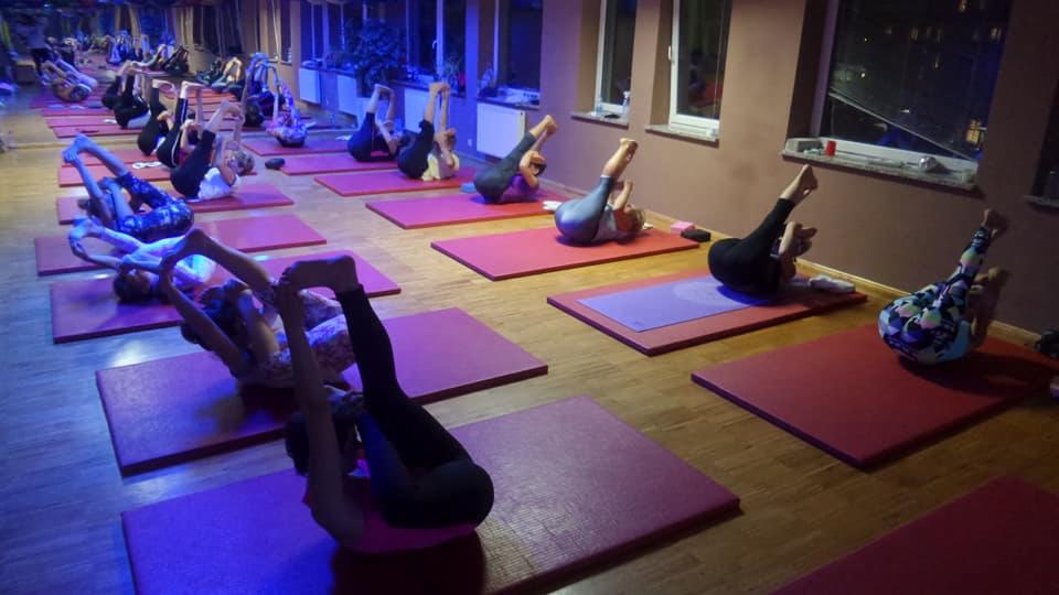 Grupa osób ćwiczy na matach w sali, wykonując pozycję z uniesionymi nogami i rękami skierowanymi ku stopom, oświetlenie sali tworzy ciepłą atmosferę.