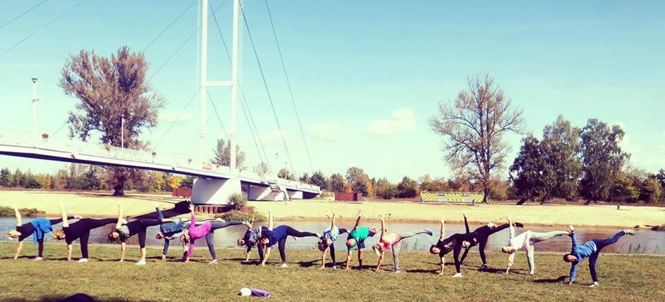 Grupa osób ćwiczy na trawie, wykonując pozycję stojącą na jednej nodze z wyciągniętą ręką, w tle rzeka i most wiszący.