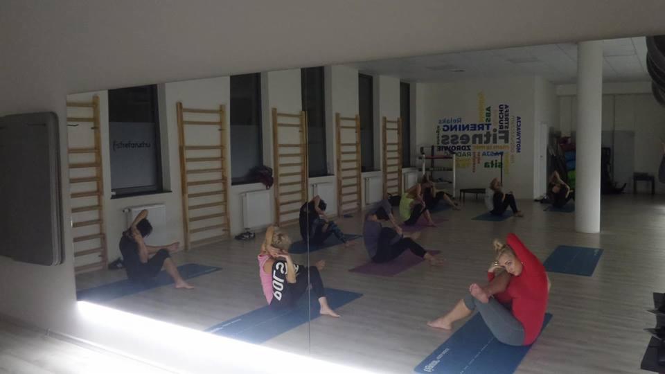 Grupa osób ćwiczy na matach w sali fitness z drabinkami gimnastycznymi i lustrami na ścianie.