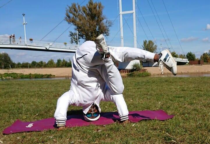 Osoba w białym dresie wykonuje stój na głowie na fioletowej macie na trawie, z nogami uniesionymi do góry, w tle most i rzeka.