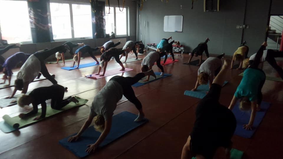 Grupa osób ćwiczy jogę w przestronnym, jasnym studio z drewnianą podłogą, wykonując pozycję psa z uniesioną nogą na kolorowych matach do ćwiczeń.