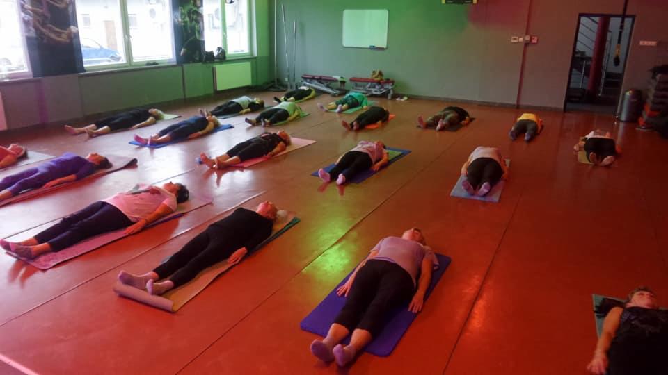 Grupa osób leżących na matach w sali gimnastycznej podczas zajęć relaksacyjnych z głowami skierowanymi w górę.