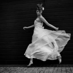 Czarno-białe zdjęcie kobiety w zwiewnej białej sukni, unoszącej się w tańcu na tle ciemnej, poziomej ściany z desek i brukowanej nawierzchni.