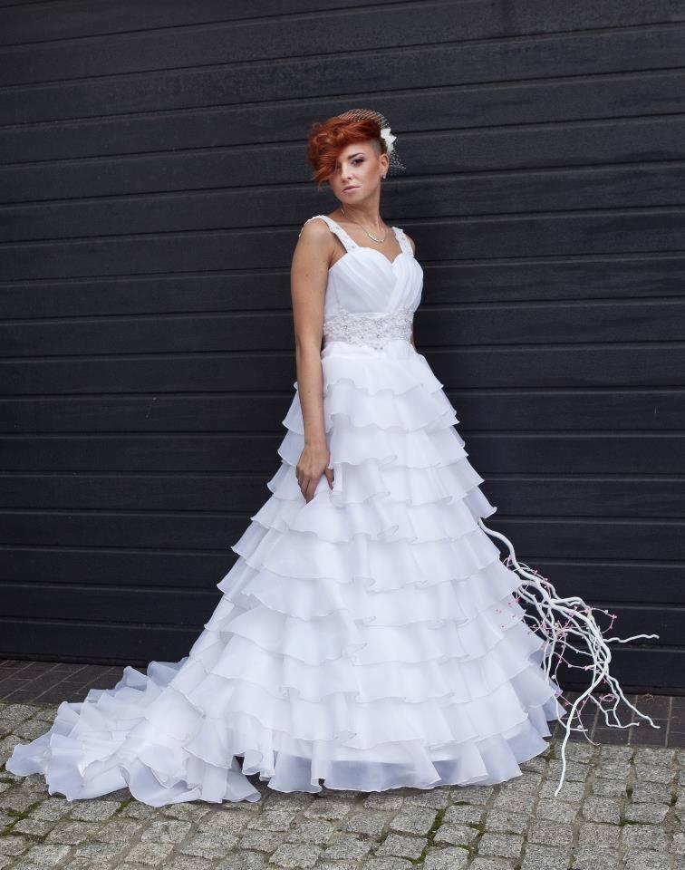 Kobieta z rudymi włosami w białej sukni ślubnej z falbanami, pozująca na tle ciemnej, poziomej okładziny ściennej i kostki brukowej.