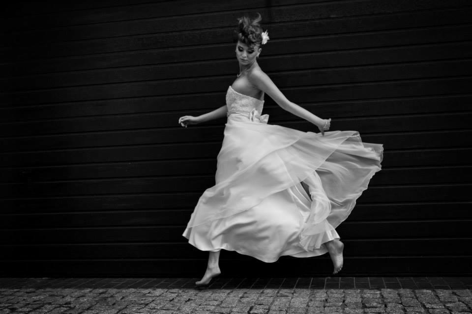 Czarno-białe zdjęcie kobiety w zwiewnej białej sukni, unoszącej się w tańcu na tle ciemnej, poziomej ściany z desek i brukowanej nawierzchni.