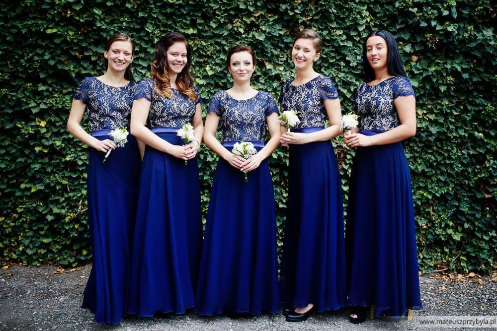 Pięć kobiet w dopasowanych, granatowych sukniach z koronkowymi topami, trzymających małe bukiety, pozujących na tle ściany z bluszczu.