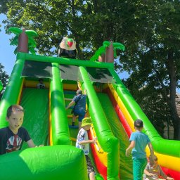 ACE ARTUR CZYŻYŃSKI - Dzieci w różnym wieku bawią się na dmuchanej zjeżdżalni w kolorach zieleni, żółci i czerwieni, stylizowanej na tropikalną wyspę z palmami i figurą tukana.