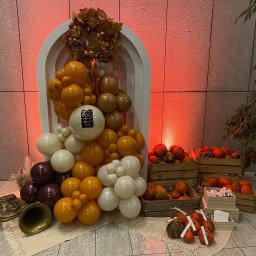 ACE ARTUR CZYŻYŃSKI - Jesienna dekoracja eventowa: kompozycja z białych i pomarańczowych balonów, dynie w drewnianych skrzynkach, wieniec z liści i trąbka na beżowym dywaniku.