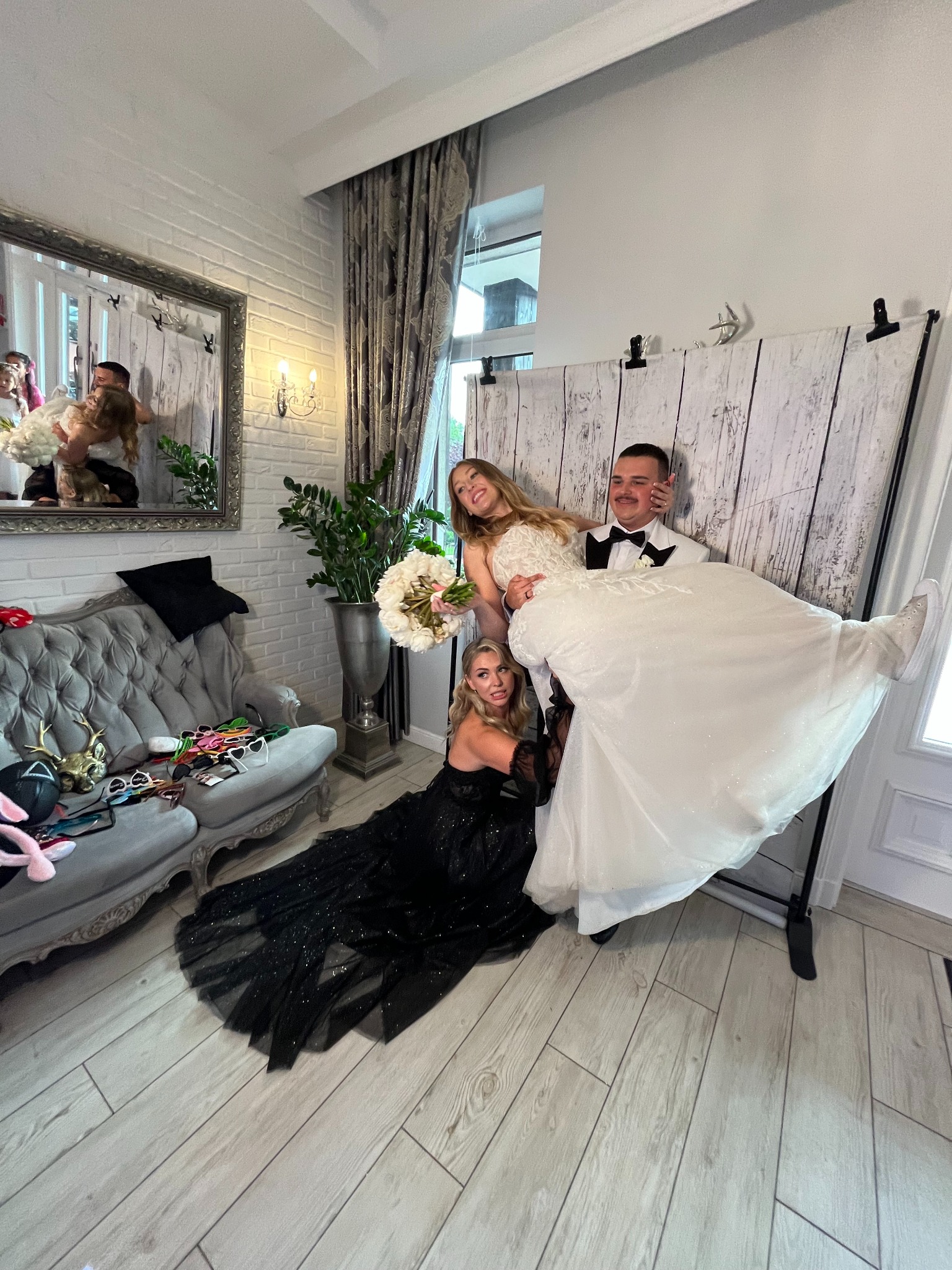 Wesołe zdjęcie ślubne w fotobudce: Panna Młoda w sukni i sneakersach, Pan Młody w smokingu, druhna w czarnej sukni. Tło z desek, rekwizyty na kanapie.