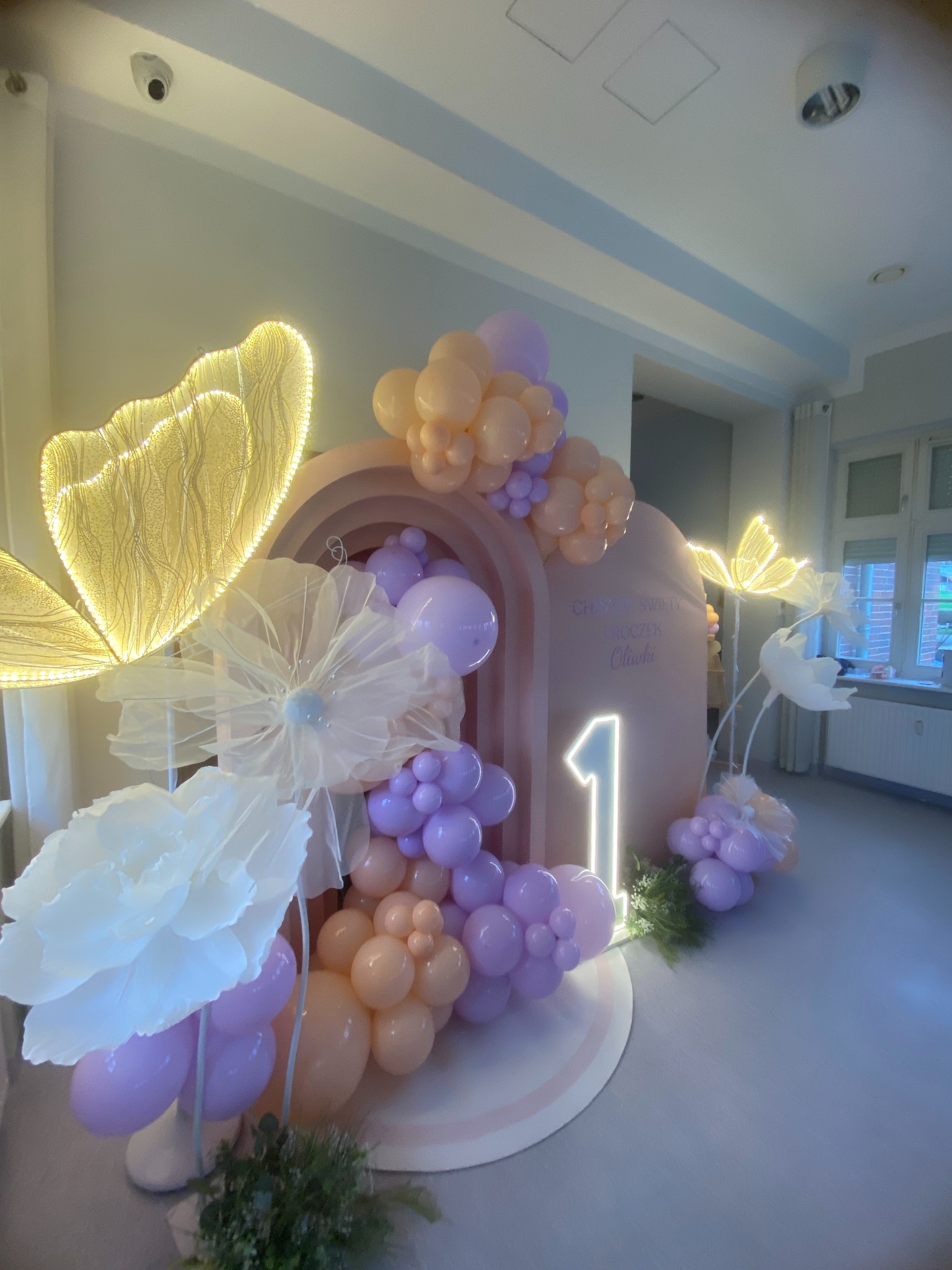Dekoracja balonowa na roczek z podświetlanymi motylami i kwiatami w pastelowych odcieniach fioletu i brzoskwini, z napisem 'Chrzest Święty Roczek Oliwki'.