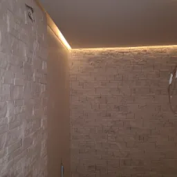 Fragment wnętrza z dekoracyjną ścianą z imitacji cegły, podświetlonej listwą LED przy suficie, sąsiadującej z gładką, jasną ścianą.