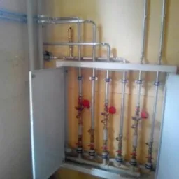 Rozdzielacz instalacji z rzędami pionowych rur z zaworami, umieszczony w szafce z otwartymi drzwiczkami, na tle ściany w odcieniach beżu.