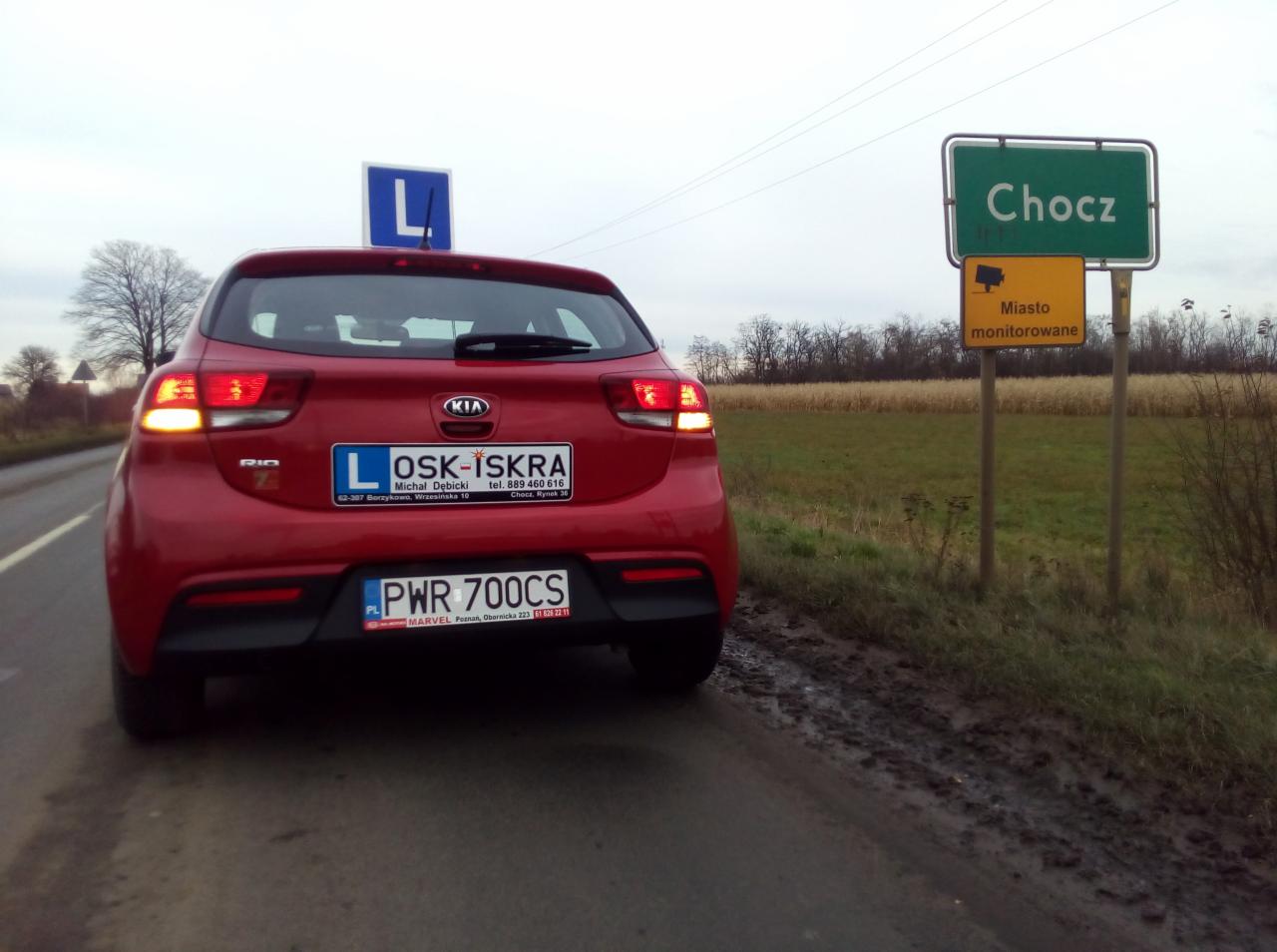 Czerwony samochód z oznaczeniem 'L' na dachu i tablicą rejestracyjną szkoły kierowców 'OSK-ISKRA' na tle znaku miejscowości Chocz i tablicy 'Miasto monitorowane'.