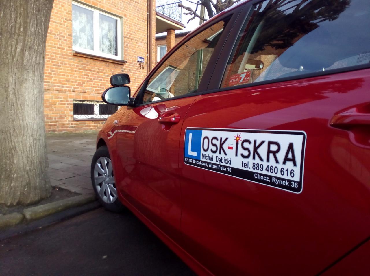 Czerwony samochód z naklejką 'OSK-ISKRA' z danymi kontaktowymi na drzwiach, zaparkowany przy chodniku i ceglanym budynku z numerem 36.