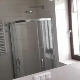 Nowoczesna łazienka z kabiną prysznicową, widoczna w odbiciu lustra, z białymi płytkami w kwiatowe wzory, oświetleniem z trzema kulistymi lampami i chromowanymi akcesoriami.