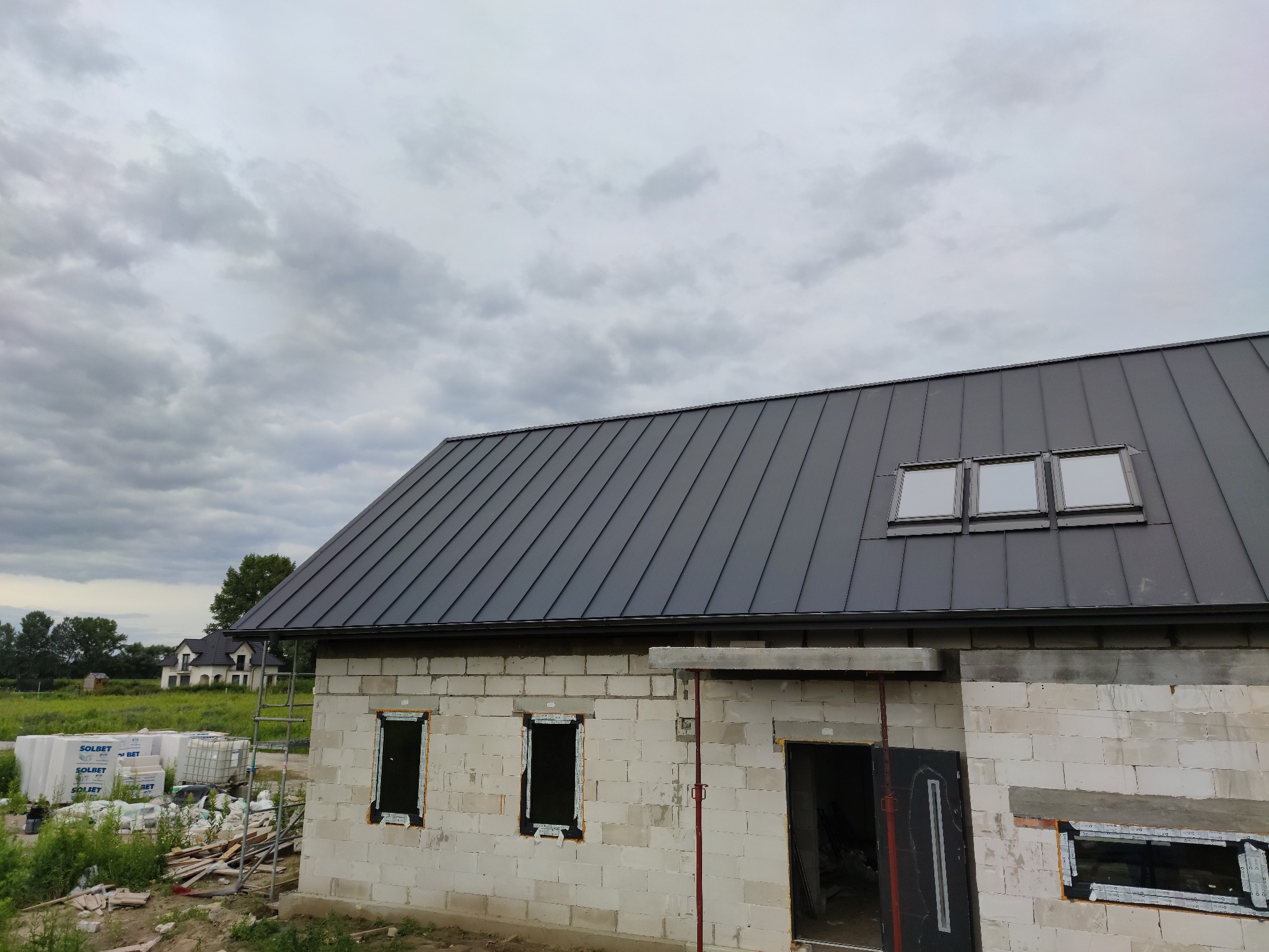 Surowy stan domu z białego bloczka z zamontowaną, grafitową blachą na dachu i oknami dachowymi, widoczne materiały budowlane i drabina na zewnątrz.