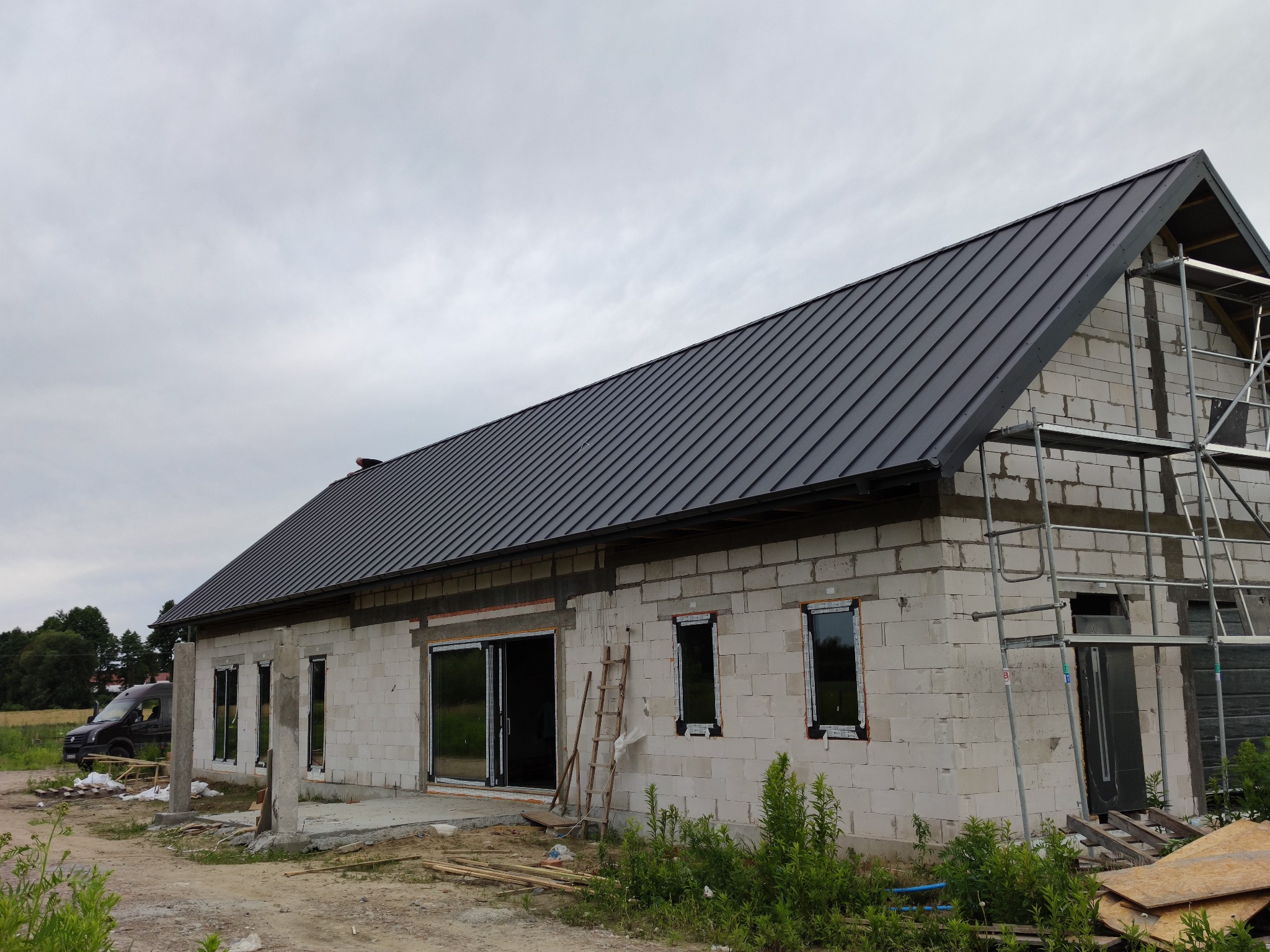 Surowy stan domu z białych bloczków z zamontowanymi oknami i grafitowym dachem z blachy na tle pochmurnego nieba, widoczne rusztowania i drabina.