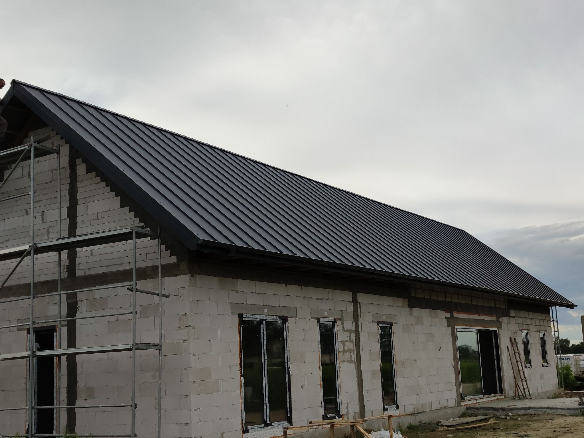 Dom w trakcie budowy z czarnym dachem z blachy na rąbek stojący, widoczne rusztowania i okna wstawione w surowy mur z bloczków.