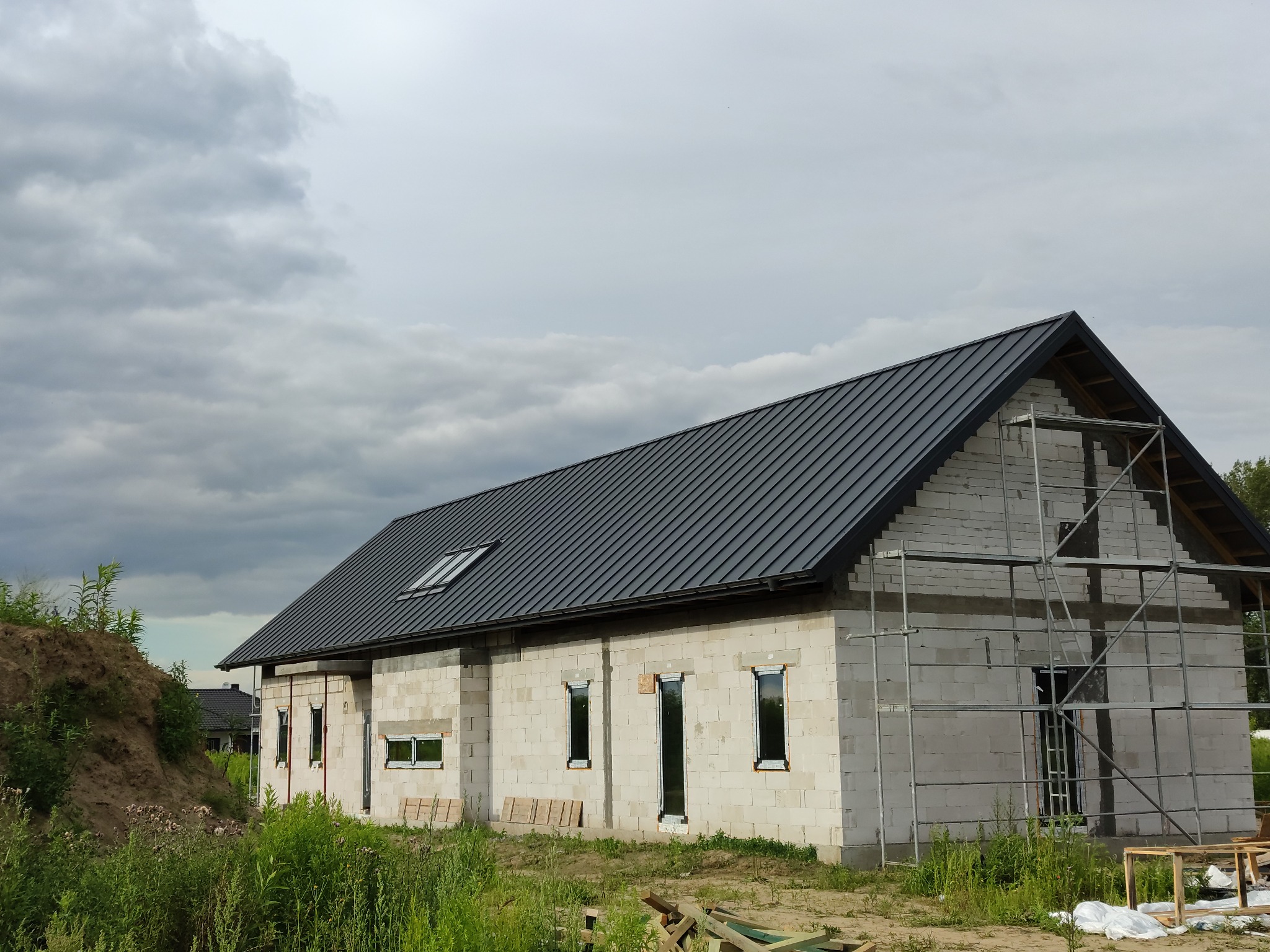 Niedokończony dom z białych bloczków z zamontowanym czarnym dachem z blachy, widoczne rusztowania, okna bez parapetów, oraz hałda ziemi z przodu.